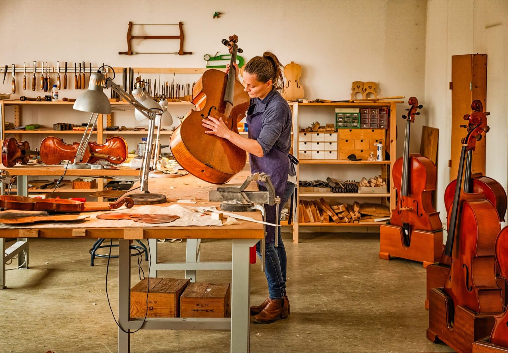 Innenaufnahme von Judith Bauers Werkstatt: Sie steht an einem Holztisch und arbeitet an einem Cello. Weitere Instrumente stehen um sie herum.